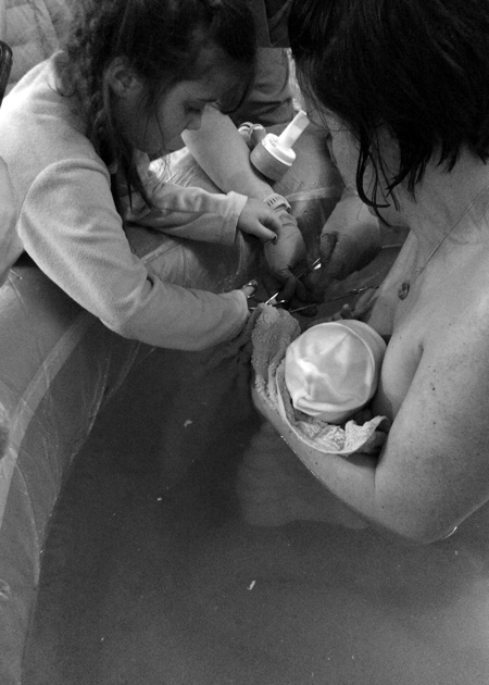 woman in birthing pool holding new born toddler cutting cord